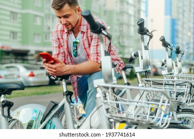 Positive man stands by the parking lot with a bicycle rental and uses a smartphone with a smile on his face. Tourist shares a bicycle for a walk on the Internet with a smartphone - Powered by Shutterstock