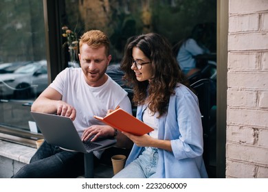 Positive man and woman in casual clothes looking at netbook screen and writing notes while sitting at small table in street cafe