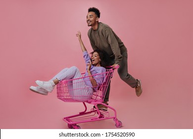 Positiver Mann und Frau mit Spaß und Reiten Einkaufswagen auf rosafarbenem Hintergrund. Fröhlicher Kerl in braunem Pullover und Hose, die sich mit der Freundin auf isoliert