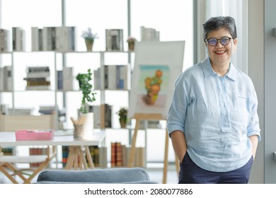 Positive Mature Asian Woman With Hands In Pockets Smiling And Looking At Camera On Blurred Background Of Home Studio