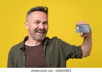 Positive Mature Beard Man Show Credit Card In Hand Isolated Over Yellow Color Background. Cool Smiling Happy Man 50s Holds Credit Bank Card, Studio Portrait. People Lifestyle Concept