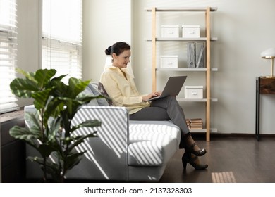 Positive mature business lady sitting on couch in office, keeping laptop on lap, typing, chatting online, watching video content, smiling. Mid adult businesswoman working in corporate lounge zone - Powered by Shutterstock