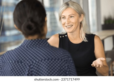 Positive mature businesswoman speaking to younger colleague in office coworking space, smiling, laughing, talking to employee, worker, giving advice for work on project - Powered by Shutterstock