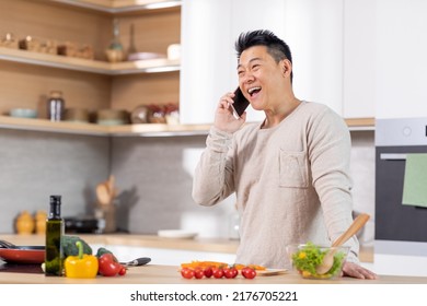 Positive Mature Chinese Man In Casual Outfit Standing Next To Kitchen Table At Home, Cooking Healthy Vegetable Salad And Talking On Mobile Phone, Laughing And Looking At Copy Space