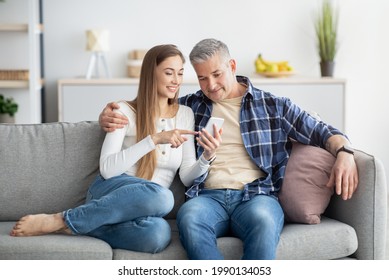 Positive Mature Couple Using Cellphone While Sitting On Couch At Home. Loving Middle-aged Family Watching Movie, Browsing Web, Having Online Meeting On Mobile Device, Indoors