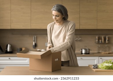 Positive mature customer woman unboxing parcel, opening package, unpacking cardboard box, at kitchen table, looking inside, receiving purchase from Internet shop - Powered by Shutterstock