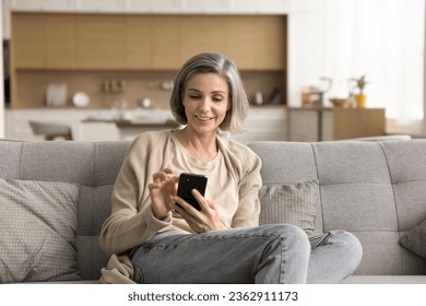 Positive mature grey haired woman enjoying online communication, domestic Internet technology at home, resting on sofa, holding mobile phone, using application for shopping, texting message - Powered by Shutterstock