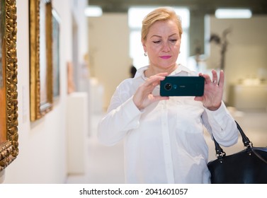 Positive Mature Lady Visitor With Mobile Phone Standing In Museum At Painting Exhibition