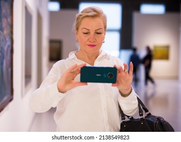 Positive Mature Lady Visitor With Mobile Phone Standing In Museum At Painting Exhibition