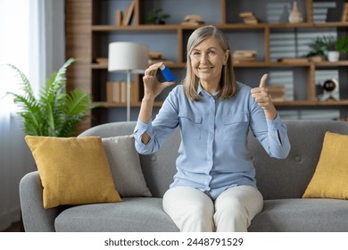 A positive mature woman comfortably sitting on a sofa at home uses an asthma inhaler. She gives a thumbs up, indicating ease and relief after use. - Powered by Shutterstock