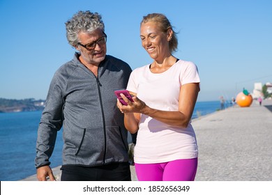 Positive Mature Woman Using Fitness App On Cell Phone After Jogging, Showing Screen To Man. Communication And Gadget For Sport Concept