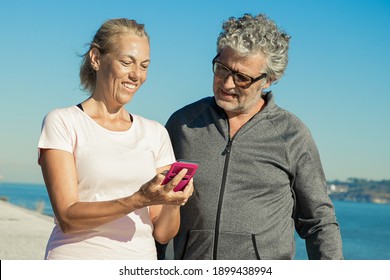 Positive Mature Woman Using Fitness App On Cell Phone After Jogging, Showing Screen To Man. Communication And Gadget For Sport Concept