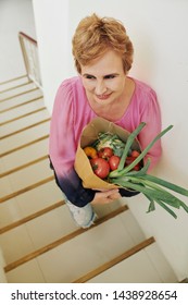 Positive Mature Woman Walking Up The Stairs And Carrying Big Package Of Groceries, View From Above