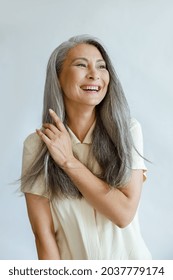 Positive middle aged Asian woman touches natural hoary hair poses for camera on light grey background in studio. Mature beauty lifestyle