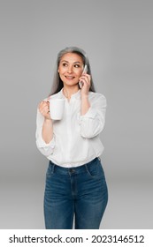 Positive Middle Aged Woman Holding Cup Of Tea While Talking On Mobile Phone Isolated On Grey