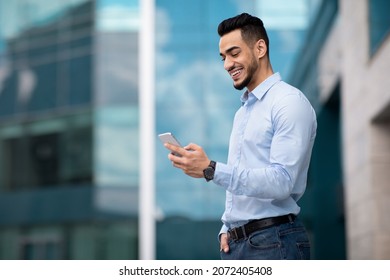 Positive middle eastern guy in formal outfit standing by modern business center, using smartphone and smiling, sending emails while having break, panorama with copy space, side view - Shutterstock ID 2072405408
