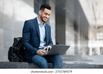 Positive middle eastern guy sitting on the street, using laptop