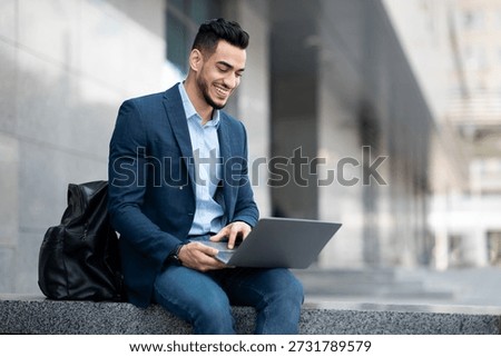 Positive middle eastern guy sitting on the street, using laptop