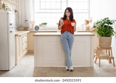 Positive middle eastern woman drinking coffee, using smartphone, standing next to kitchen counter, copy space - Powered by Shutterstock