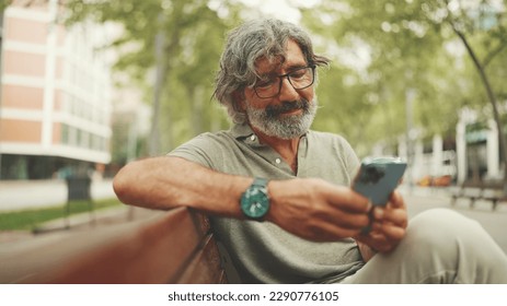 Positive middle-aged man with gray hair and beard wearing casual clothes sits on bench and uses mobile phone. Mature gentleman in eyeglasses writes message on smartphone - Powered by Shutterstock