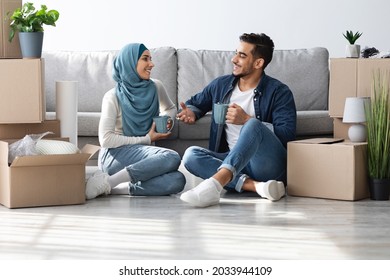 Positive middle-eastern young family husband and wife in hijab sitting on floor in empty room, drinking coffee while unpacking, having conversation and smiling, planning house renovation - Powered by Shutterstock