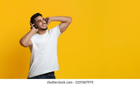 Positive millennial arabic guy in white t-shirt using brand new wireless stereo headset, listening to music over yellow studio background, looking at copy space and smiling, panorama