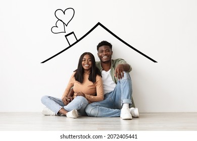 Positive millennial black couple sitting under roof illustrated on wall at their new apartment, hugging and smiling at camera. Happy african american family buying new house, creative image