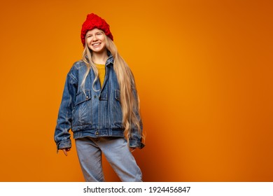 Positive Millennial Hipster Female Model In Denim Jacket And Red Knitted Hat Looking At Camera Against Bright Orange Background