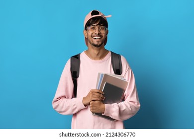 Positive Millennial Indian Guy Student In Pink Baseball Cap And Sweatshirt Posing On Blue Background, Holding Books And Notepads, Carrying Backpack, Copy Space. Education, School, College Concept