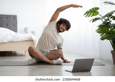 Positive Millennial Indian Guy In T-shirt And Shorts Sitting On Yoga Mat, Looking At Laptop Screen And Stretching, Having Fitness Class Online In The Morning, Bedroom Interior, Copy Space