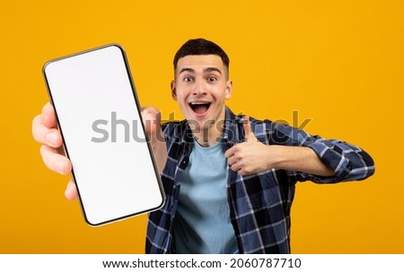 Positive millennial man showing huge brand new smartphone with empty screen and thumb up over yellow studio background, mockup, panorama with copy space, newest mobile app concept