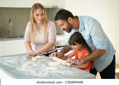 Positive Mom And Dad Teaching Daughter To Roll Dough On Kitchen Table With Flour Messy. Young Couple And Their Girl Baking Buns Or Pies Together. Family Cooking Concept