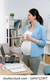 Positive Mood Lifestyle. Straight Photo Side View Happy Asian Korean Pregnant Woman Drinking Cup Of Tea While Standing At Work Desk With Laptop In Cozy Home Office. Elegant Maternity Female Worker