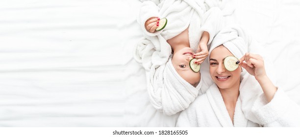 Positive mother and daughter doing face mask applying pieces of cucumber to their eyes, wrapped in a white bath towel, Facial skin care, cosmetology and spa, Long banner format. space for text. - Powered by Shutterstock