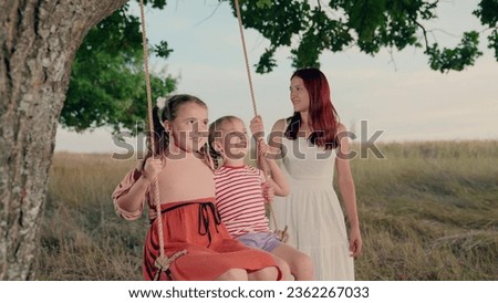 Positive mother rides cute little girls on swings in summer park at twilight time. Little girls play swings with loving mother in country field. Mother amuses little daughter on swings in garden