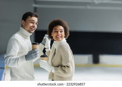 Positive multiethnic couple holding hands and coffee to go on ice rink  - Shutterstock ID 2232926857