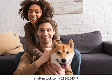 Positive multiethnic couple looking at camera near blurred shiba inu in living room - Powered by Shutterstock