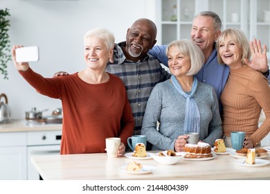 Positive multiethnic senior friends taking selfie on cell phone at kitchen, happy good-looking elderly men and women drinking tea, eating cake together, having home party, enjoying time together - Powered by Shutterstock