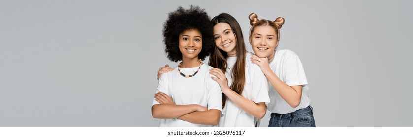 Positive multiethnic teen girlfriends in casual t-shirts and jeans hugging each other and looking at camera together isolated on grey, lively teenage girls concept, banner with copy space - Powered by Shutterstock