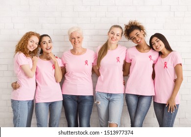 Positive Multiethnic Women In Breast Cancer T-Shirts With Pink Ribbons Hugging Standing Over White Brick Wall Indoor.