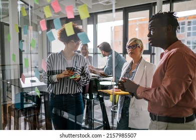 Positive Multiracial Group Of Business People Working Together On A New Project And Writing Main Topics And Sharing Creative Ideas While Using Colorful Sticky Notes On The Glass Wall.