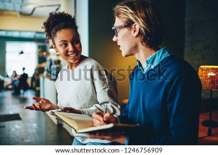 Positive multiracial male and female students having conversation about common project in cafe making notes, man writing to do list in notepad while talking and discussing with his girlfriend