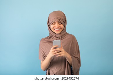 Positive Muslim Teen Girl In Hijab Using Cellphone, Checking Messages, Browsing Social Media Over Blue Studio Background. Adolescent In Traditional Headscarf Chatting Online, Having Video Call