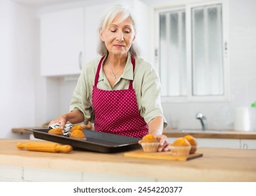 Positive old woman in apron taking out just baked cupcakes. Senior woman cooking cakes at home. - Powered by Shutterstock