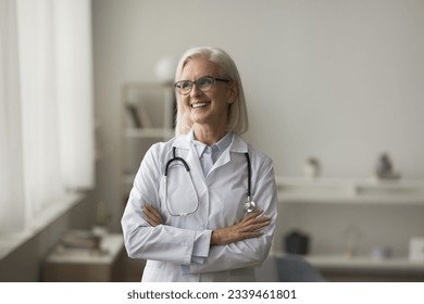Positive older mature practitioner woman in glasses posing indoors, looking at window away, thinking, smiling. Senior medical professional, doctor in white uniform casual portrait