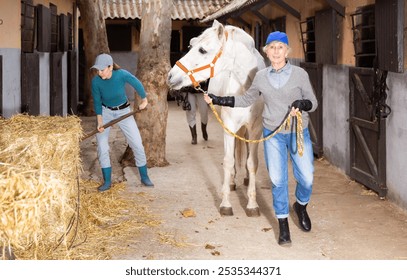 Positive older woman stable keeper leading white horse along stalls outdoor while young female worker stacking hay with pitchfork in yard. Daily work in horse farm concept - Powered by Shutterstock