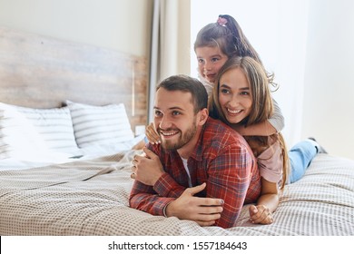 Positive Optimistic Man Lying On Bed In Comfortable Hotel Room, Keeping Hands Crossed Over, Looking Away, Wife And Child Happily Resting On His Back, Holiday Time Concept, Shot From Below