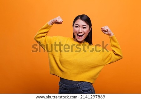 Positive optimistic woman raising arms while feeling strong and confident on orange background. Motivated triumphant girl celebrating victory and feeling optimistic and independent.