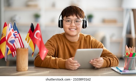 Positive Overweight Chinese Kid Teenager With Wireless Headset And Digital Tablet Sitting At Table By Various International Flags, Studying Foreign Languages On Internet, Using Movile App, Panorama