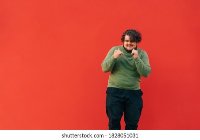 Positive Overweight Man And Green Sweatshirt Funny Dancing On Red Wall Background And Smiling. Fat Man Shows A Dance Performance. Isolated.